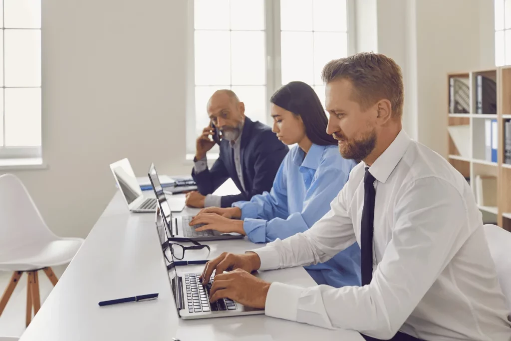 Professionals working on laptops at a long table in a bright office space.