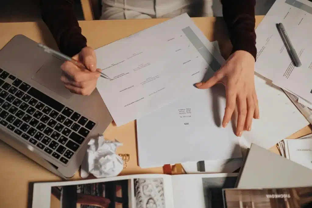 Detailed view of a person's hands organizing documents and writing notes beside a MacBook.