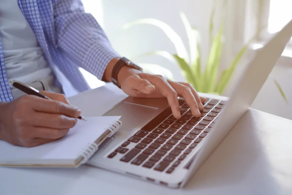 Person typing on laptop while writing notes in a notebook.