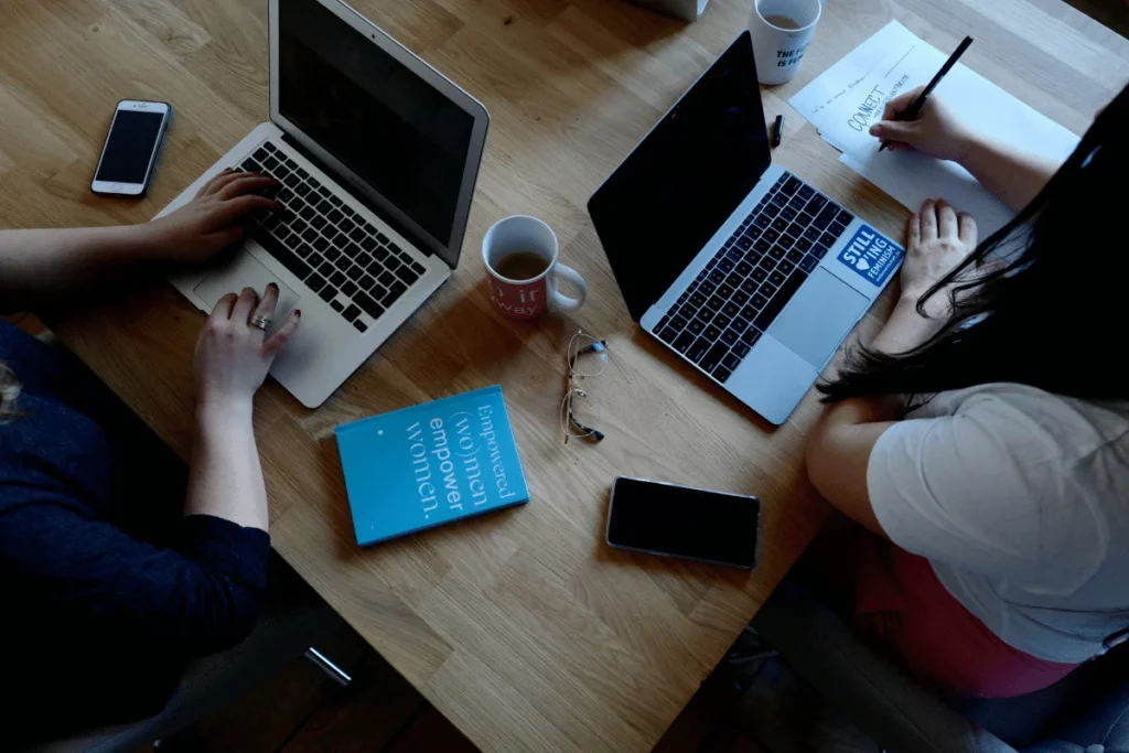 Two individuals working at a wooden table with laptops, mobile phones, a notebook, and a mug.