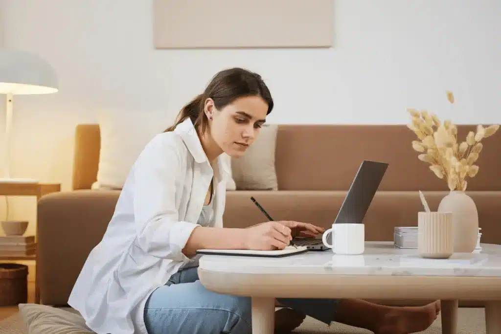Concentrated woman jotting down notes while working on a laptop at home.