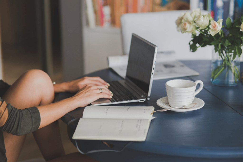 Woman typing on a laptop at a home office setup with a diary and a cup of coffee.