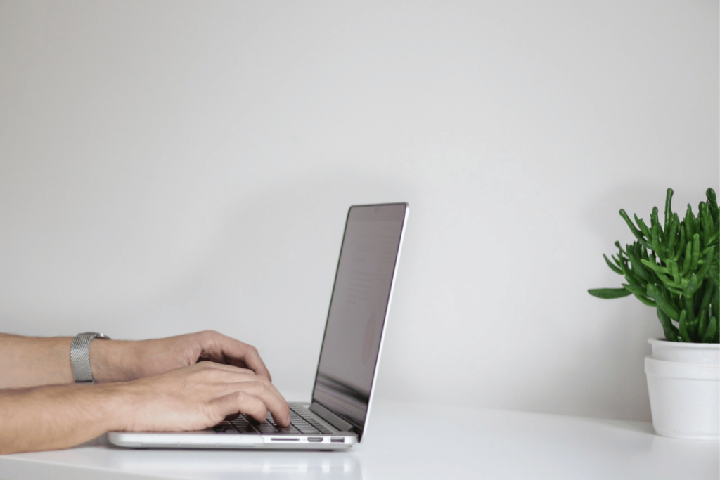 Close-up of hands typing on a modern laptop next to a potted plant.