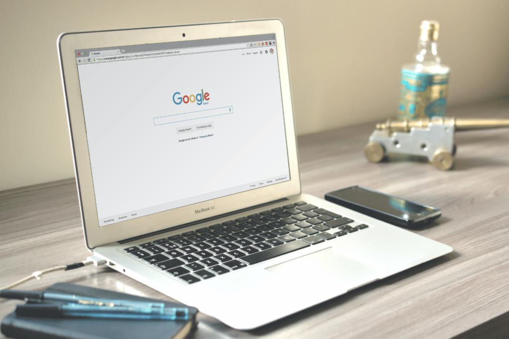 Close-up of an open MacBook with Google homepage on screen, phone and decor on wooden table.