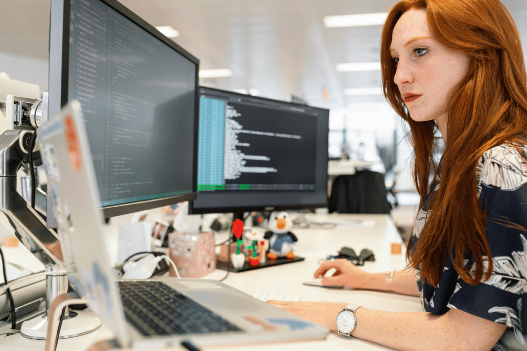 Focused female programmer coding on dual monitors in a bright office.