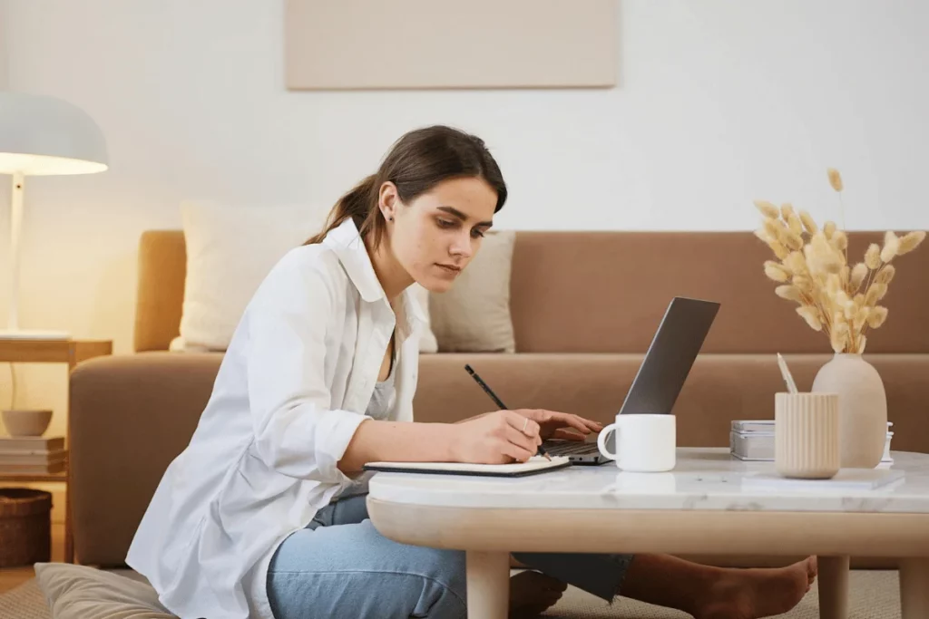 Home office setup with a woman working on a laptop and taking notes at a coffee table.