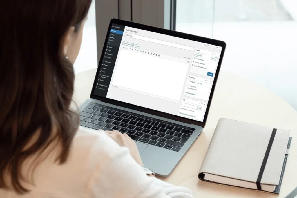 A person using a laptop to manage a blog or website, with a notepad on the side for additional notes.