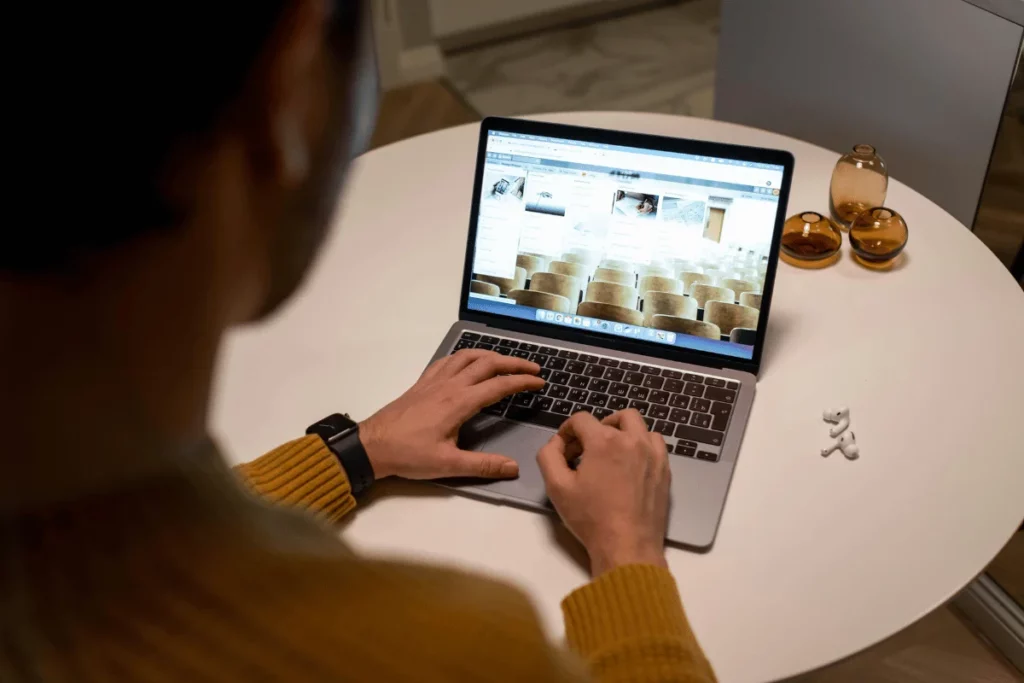 Man browsing a website on a laptop, sitting at a round white table with amber-colored decor pieces and earbuds.