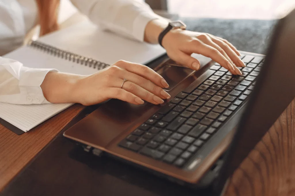Typing on a laptop while taking notes in a notebook.