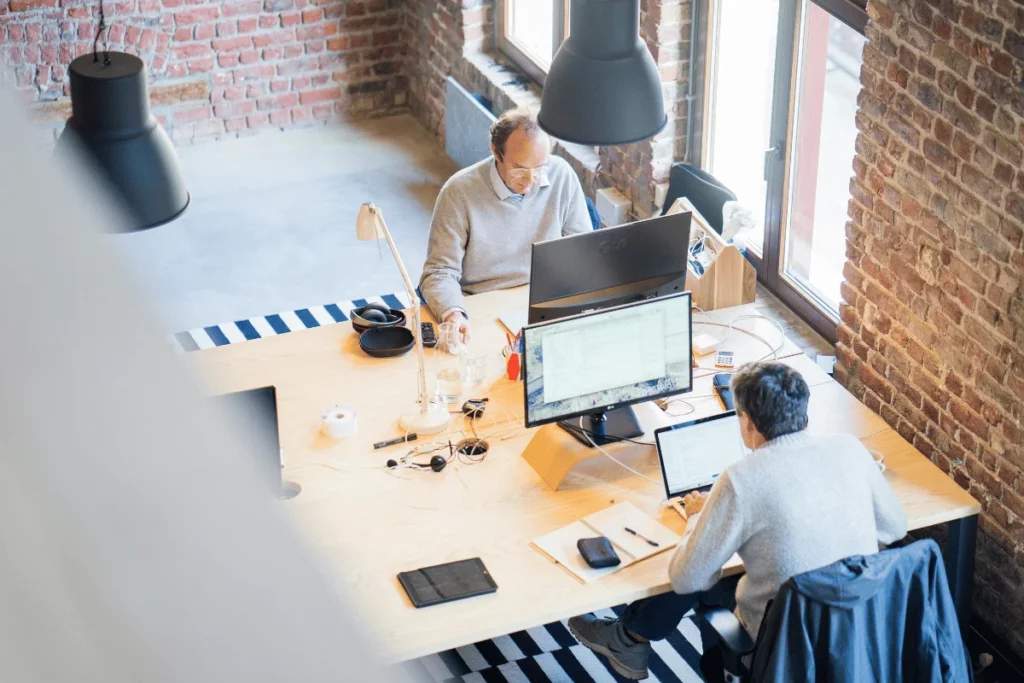 Two colleagues working on laptops and desktops in an industrial-style office.