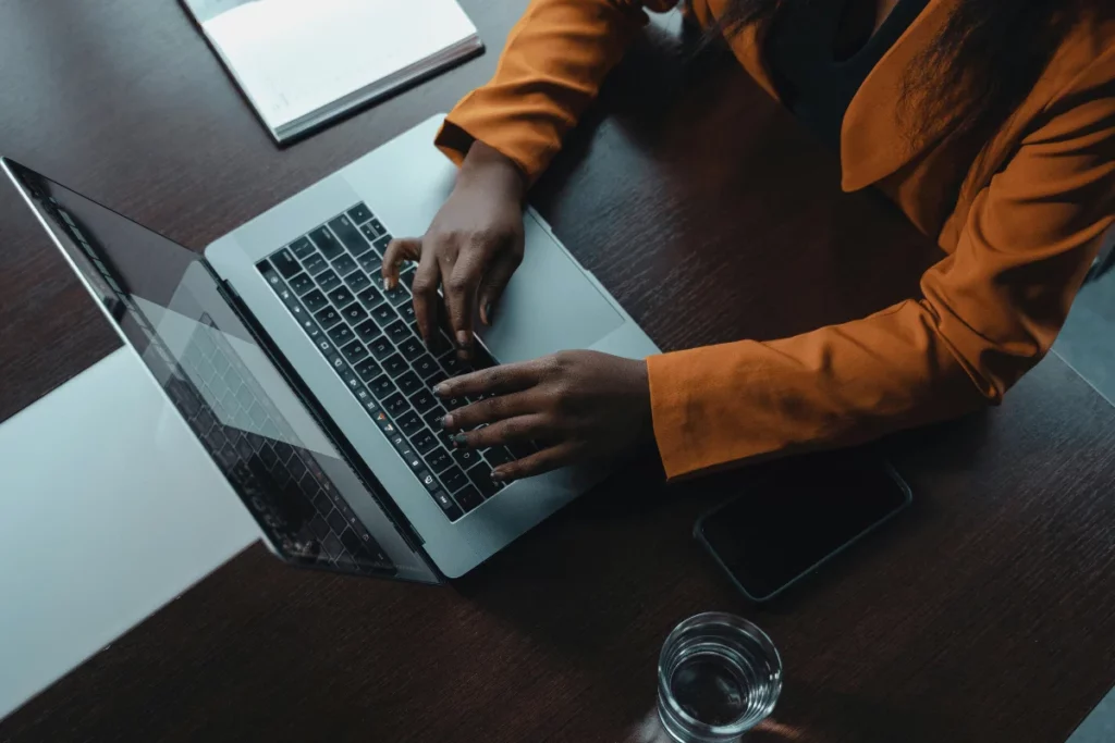 Individual in an orange blazer working on a laptop, with a smartphone and a glass of water nearby.