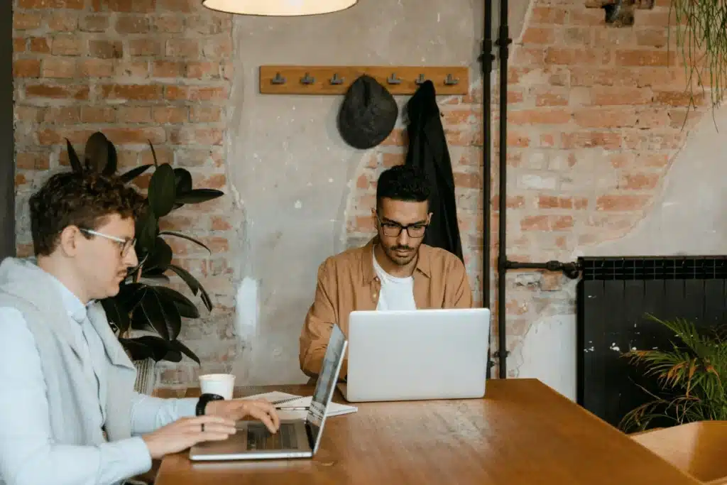 Two people are working at a wooden table in a cozy setting with exposed brick walls, laptops, and greenery around.