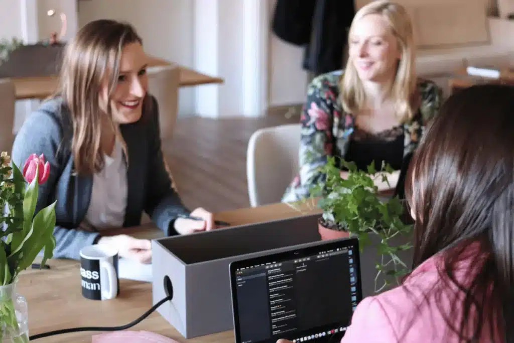 Three individuals engage in a meeting at a wooden table adorned with a potted plant and a coffee mug, with a laptop at the center.