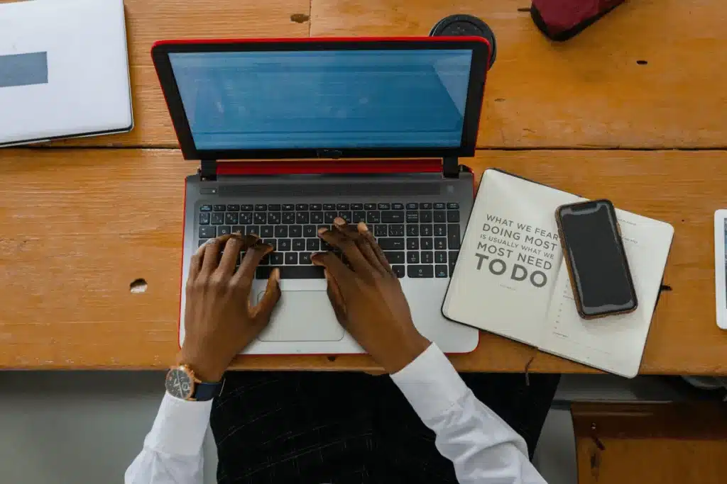 A person types on a laptop with a notebook and smartphone nearby, on a wooden table, emphasizing productivity and focus.