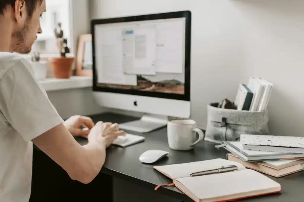 A person types on a computer keyboard, surrounded by notebooks, a coffee mug, and a cozy workspace with plants.